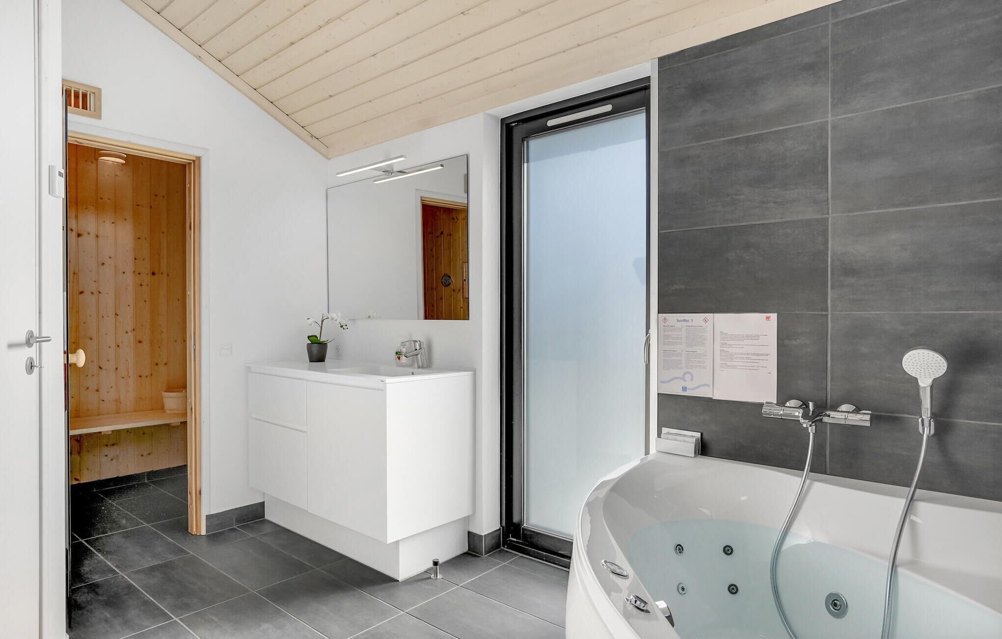 A modern bathroom with a white vanity, a glass-enclosed shower, and a wooden door leading to a sauna.