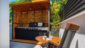 An outdoor kitchen area with a grill, a round table with chairs, and a wooden pergola. - Awesome apartment in Obrovac (Obrovac)