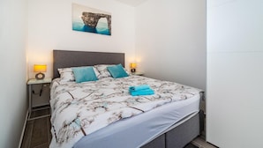 A neatly made bed with a floral bedspread, two pillows, and a rolled towel. There is a wall-mounted picture above the bed and two bedside lamps on either side. - Awesome apartment in Obrovac (Obrovac)