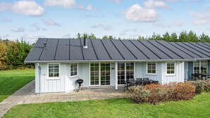 Un bâtiment blanc avec un toit foncé, de grandes fenêtres et un patio aménagé avec du mobilier d’extérieur.