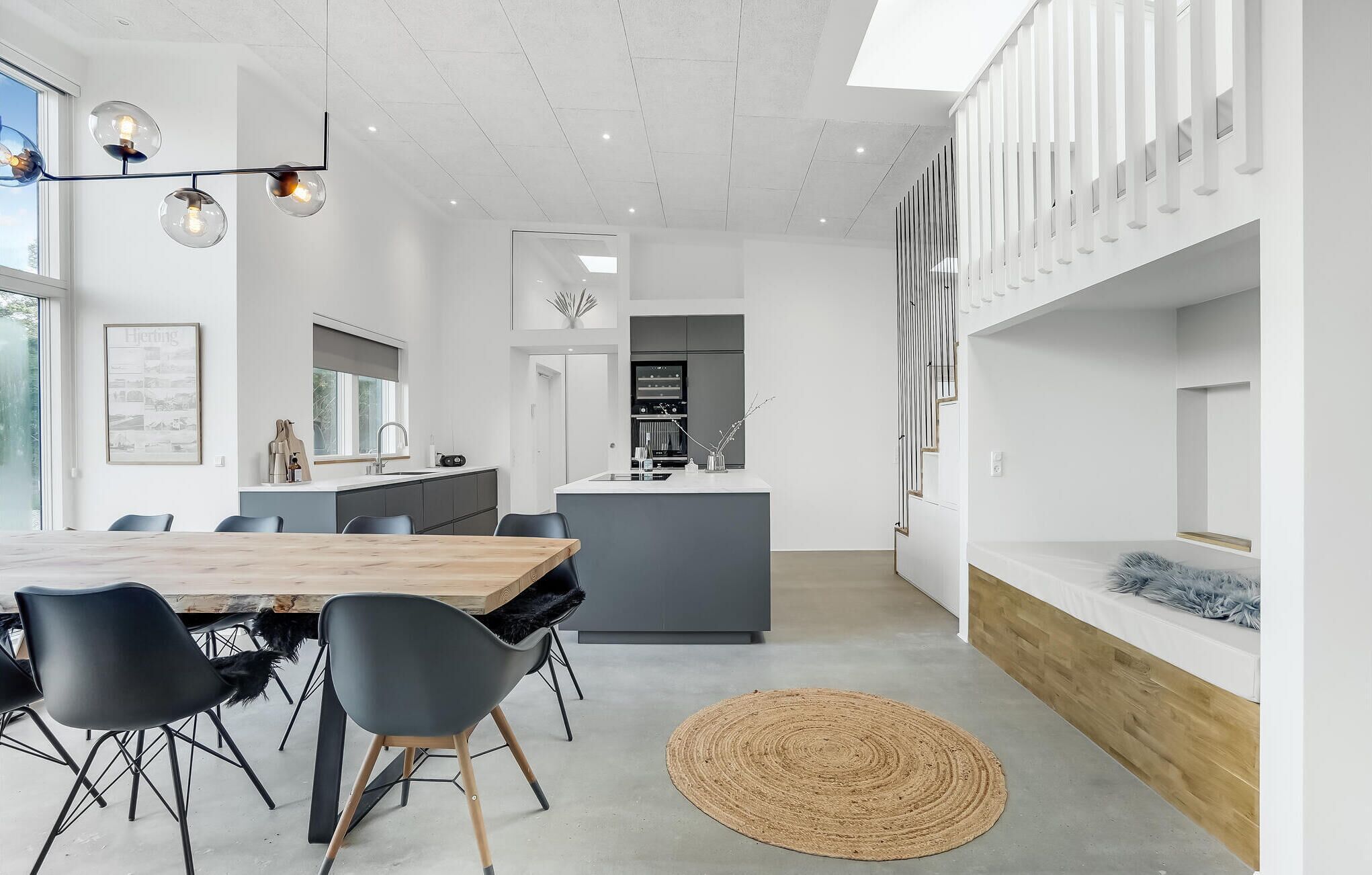 Una cocina moderna con una isla central, un área de comedor con mesa y sillas de madera, y una escalera con barandilla blanca.