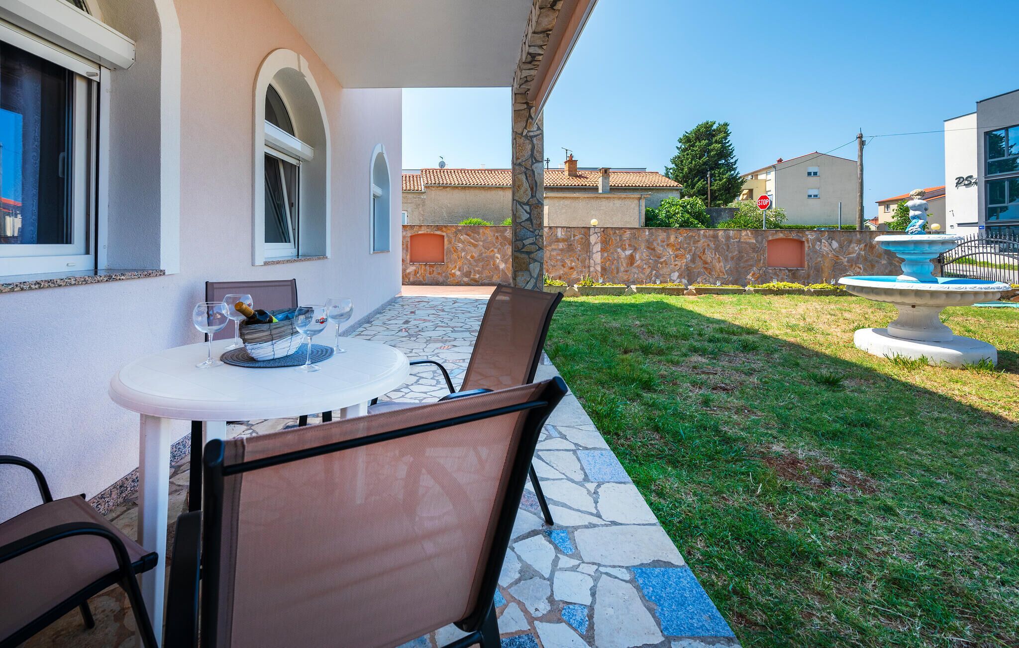 An outdoor patio with a table and chairs, a stone-tiled floor, and a view of a residential area with houses and a fountain.