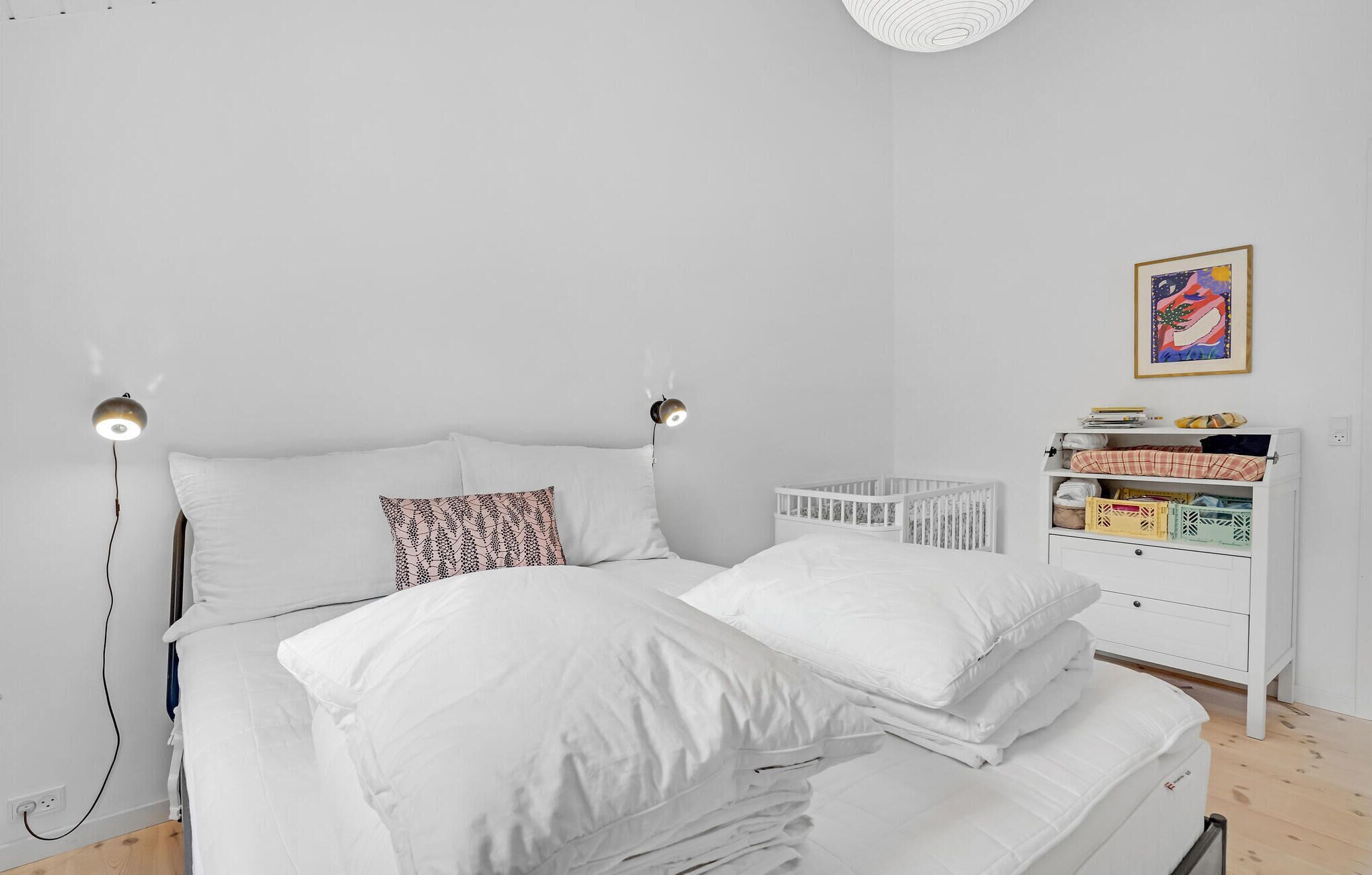 A minimalist bedroom with a white bed, two bedside lamps, a crib, and a storage unit with boxes.
