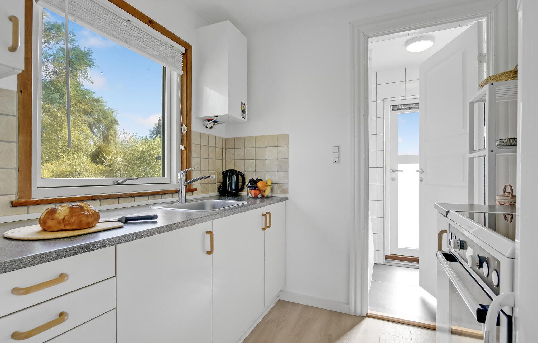 Ett modernt kök med vita skåp, ett fönster med utsikt över grönområden, en bänkskiva med ett bröd, en vattenkokare och en fruktskål.