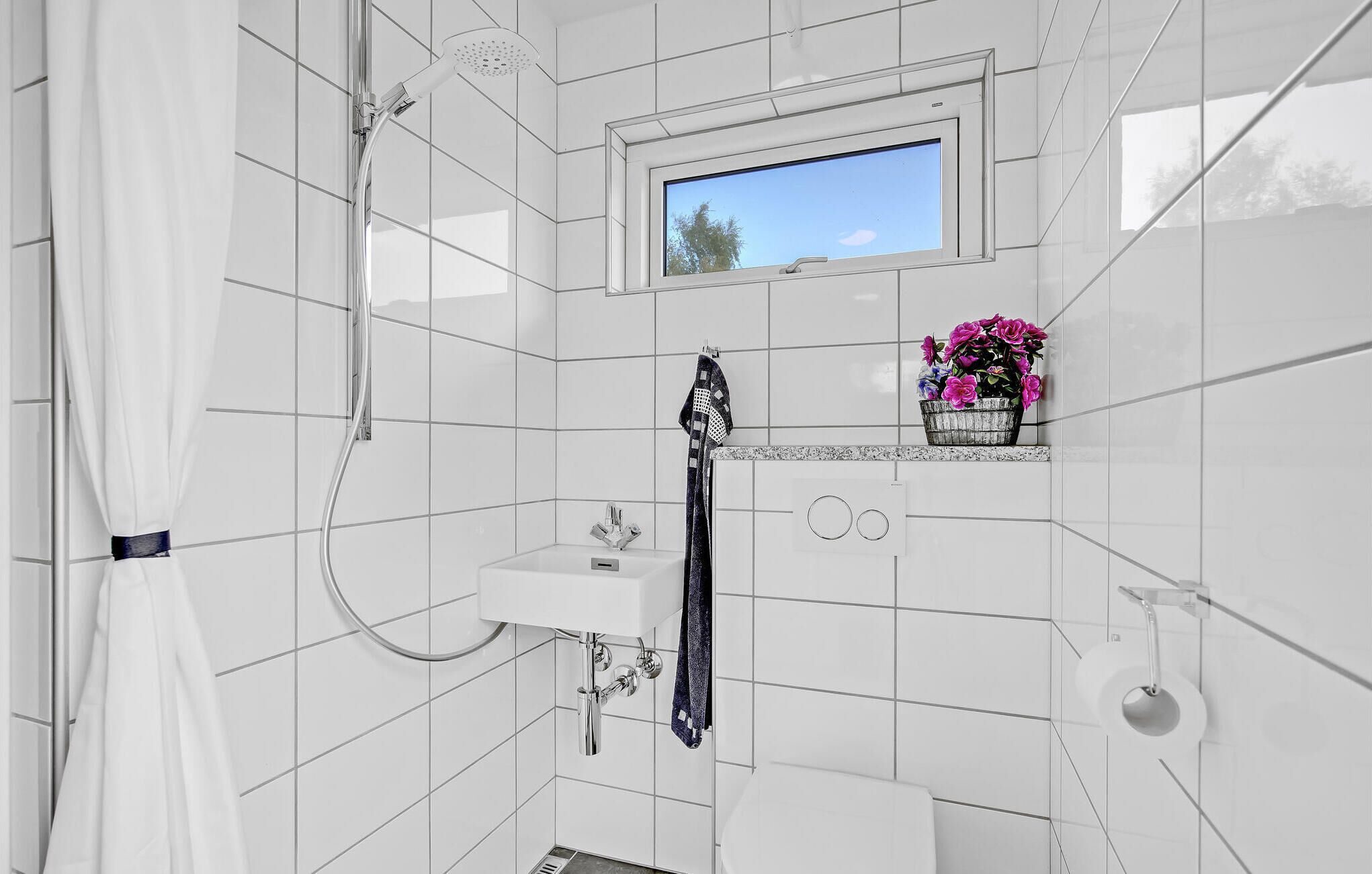 Un bagno moderno con piastrelle bianche, una doccia, una finestra e un piccolo ripiano con fiori.