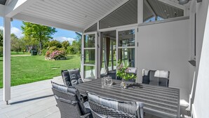 Une terrasse extérieure avec une table et des chaises, une porte vitrée donnant sur un jardin et un mur extérieur blanc.