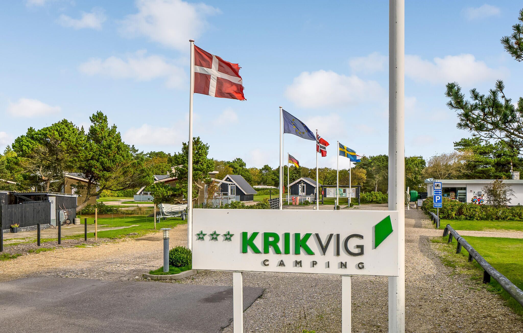 Une enseigne indiquant « KRIKVIG CAMPING », avec plusieurs drapeaux, dont ceux du Danemark, de la Suède et de l’Union européenne, et un petit bâtiment en arrière-plan.
