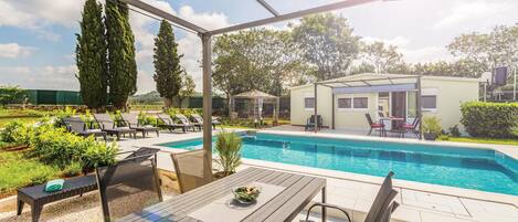 A covered outdoor area with a pool, lounge chairs, and a table with a bowl of fruit.