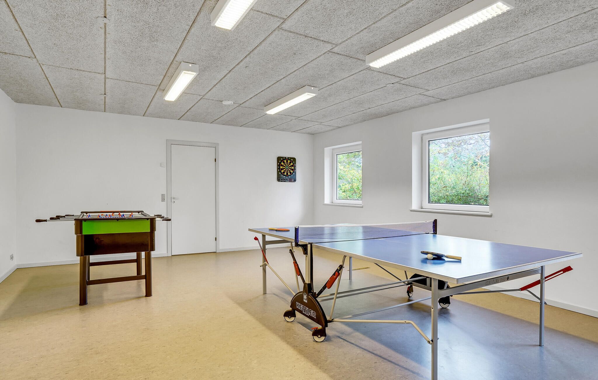A room with a ping pong table, a foosball table, and a dartboard. There are windows and a door.