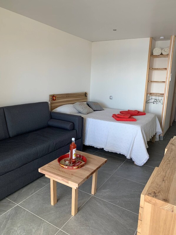 A modern, well-lit room with a dark grey sofa, a wooden coffee table with a tray, a bed with white bedding and red towels, and a wooden shelf unit against the wall.