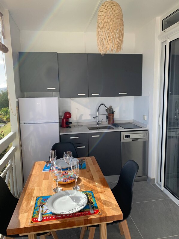 Una cocina moderna con armarios grises, un refrigerador blanco y una tetera roja. Hay una mesa de comedor con un mantel individual, vasos y un plato.