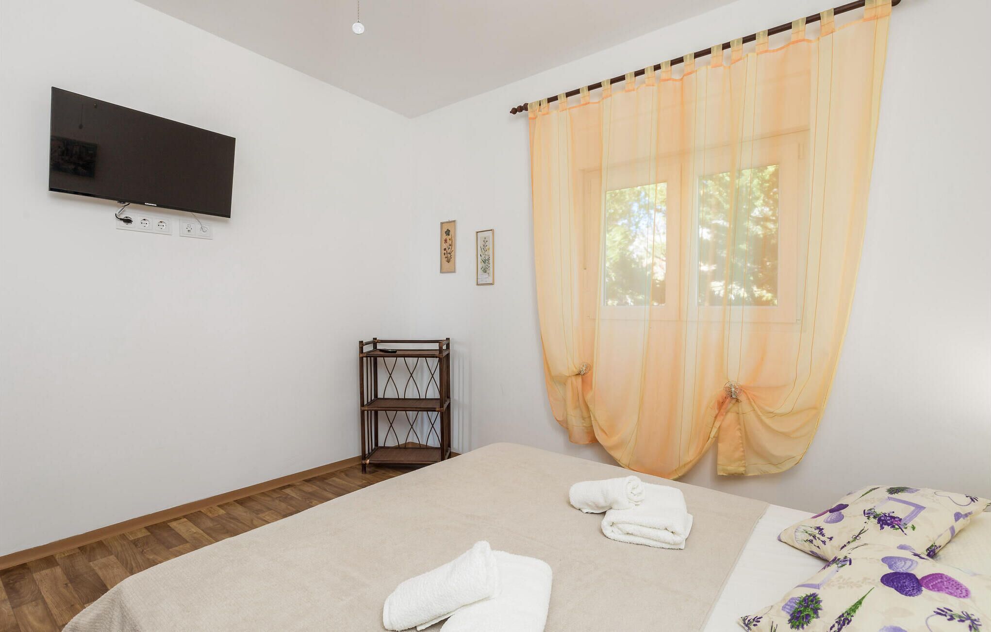 Une chambre à coucher avec un lit, un téléviseur fixé au mur, un plancher en bois et une fenêtre avec des rideaux.