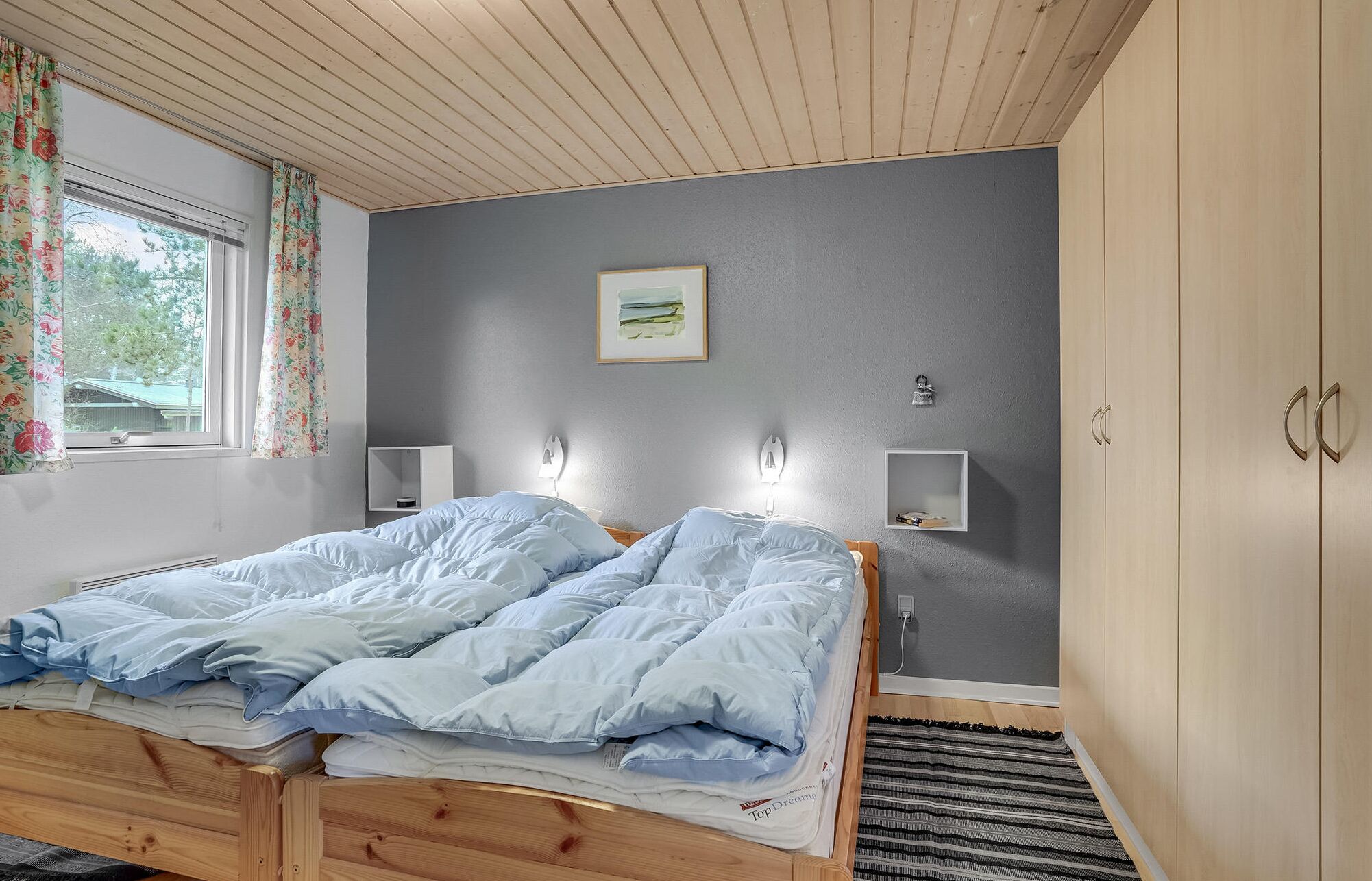 A bedroom with a wooden bed, grey walls, a window with floral curtains, a painting, and a wardrobe.