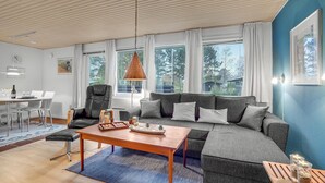En moderne stue med træloft, store vinduer, en grå sofa, et træbord og et spiseområde med et hvidt bord og stole.