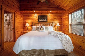 Ein Schlafzimmer einer Holzhütte mit einem großen Bett, zwei Nachttischlampen, einem gerahmten Bild an der Wand und einem Fenster mit Jalousien.
