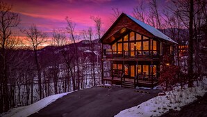 A two-story log cabin with a balcony, surrounded by snow-covered ground and trees, under a twilight sky.