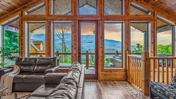 A spacious wooden interior with a large windowed door leading to an outdoor deck, leather sofas, and a view of mountains.