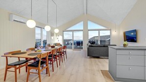 Un amplio comedor con techo alto y ventanas grandes, un sofá gris y una cocina moderna con televisión de pantalla plana.