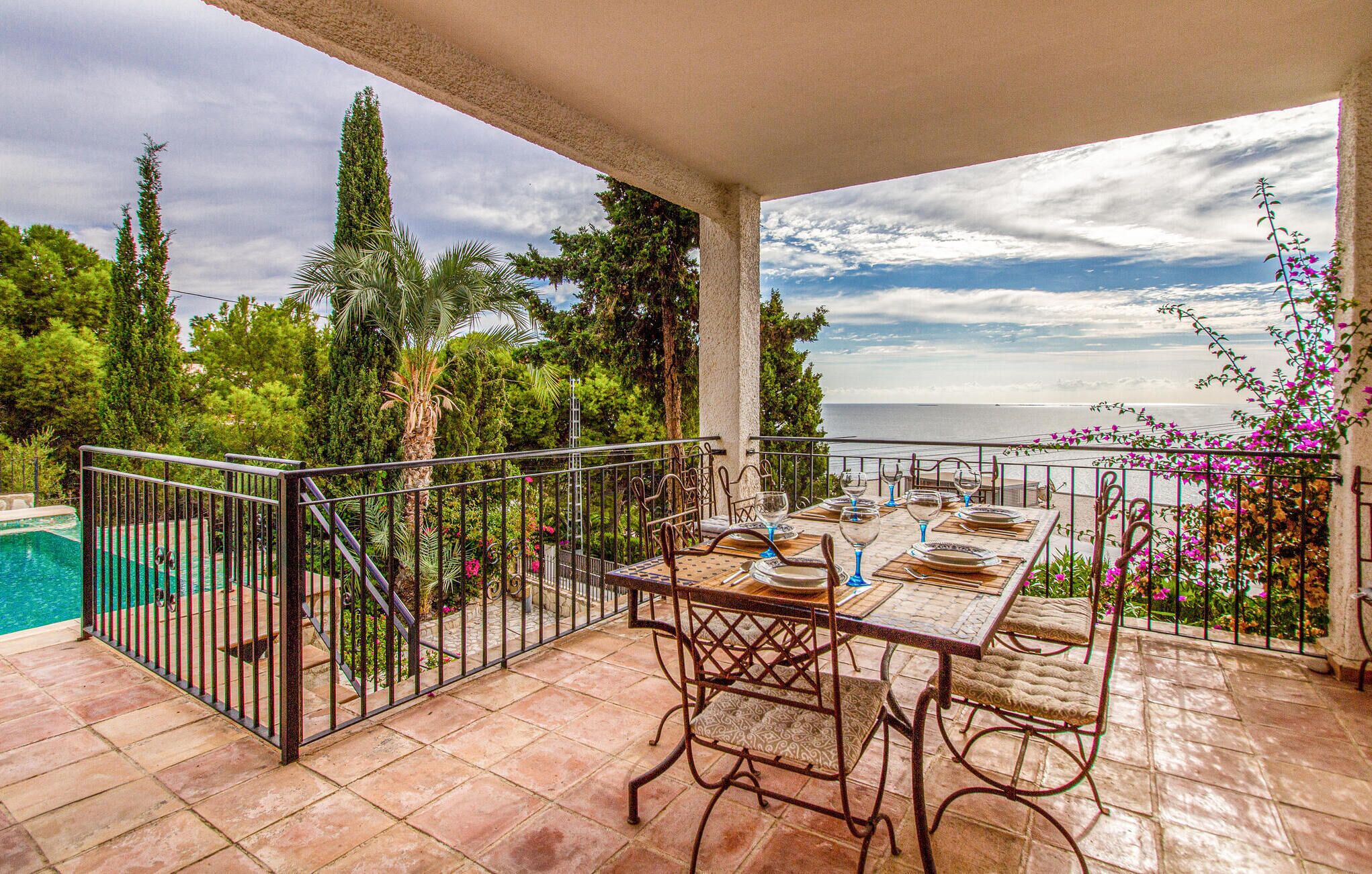 A terrace with a table set for dining, overlooking a pool and a scenic view of the sea.