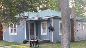 Une maison de plain-pied avec un porche, une petite table et des chaises, et un panier à fleurs suspendu.