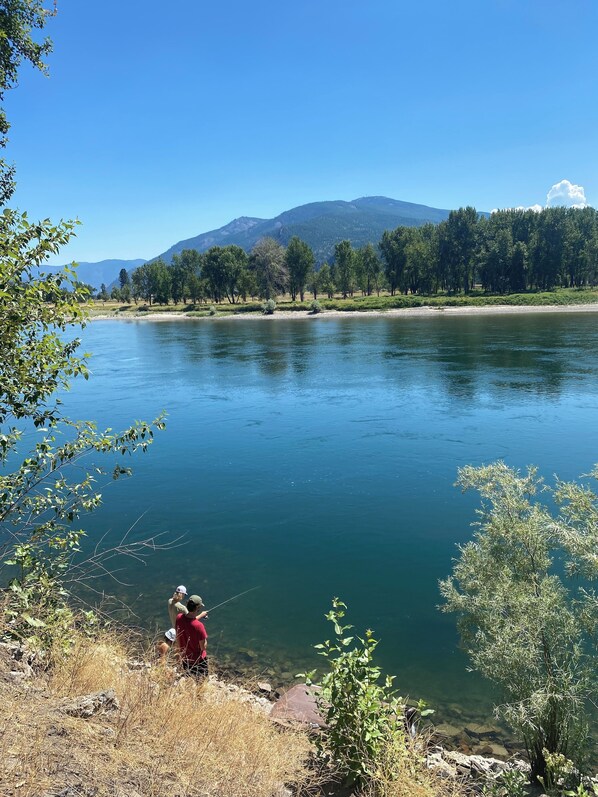 A serene riverside scene with a person fishing, surrounded by trees and mountains in the background.
