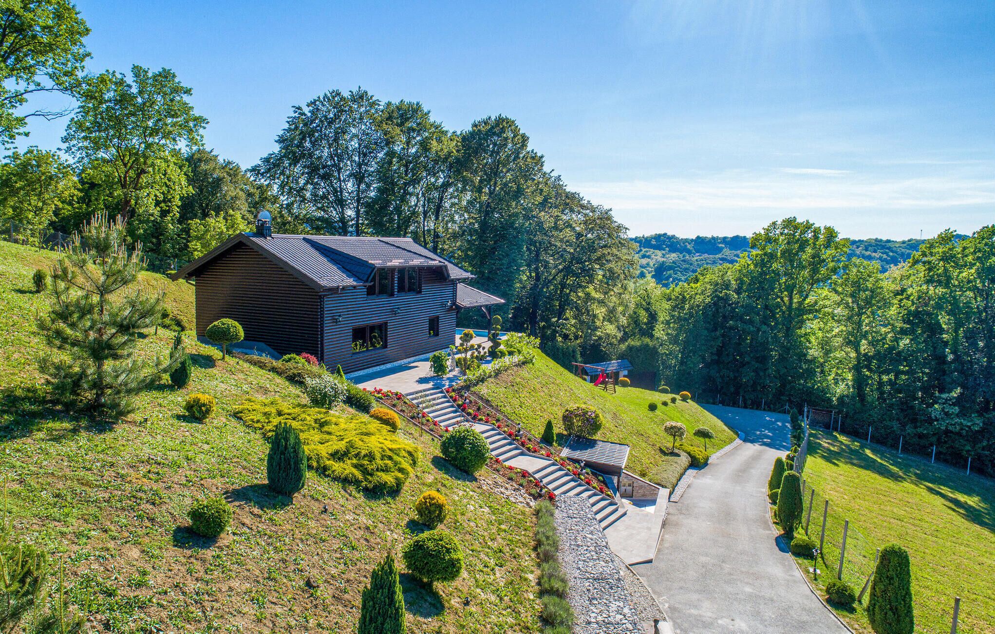A modern wooden house with a sloped roof, surrounded by a well-maintained garden and a winding driveway.