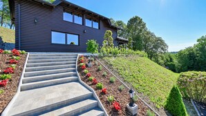 Una casa moderna con techo inclinado, una escalera de concreto que lleva a la puerta principal, y un jardín con diversas plantas y flores.