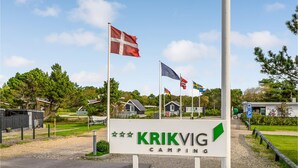 Cartell de 'Krikvig Camping' amb diverses banderes, incloent la bandera danesa, i una vista clara de l'entorn amb cases i vegetació.