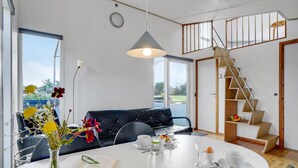 Un saló modern amb un sofà de cuir negre, una taula de dinar a punt amb un llibre, gots i un plat de menjar, i una escala que condueix a un nivell superior.