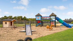 Un parc infantil amb tobogan, elements per escalar i un petit edifici de fusta.