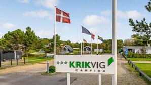 Señal de 'KRIKVIG CAMPING' con varias banderas, incluyendo las de Dinamarca, Suecia y la bandera de la Unión Europea, y un edificio pequeño al fondo.