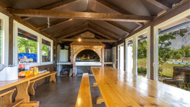 A covered outdoor kitchen area with a brick oven, wooden benches, and large windows offering views of the outdoors.