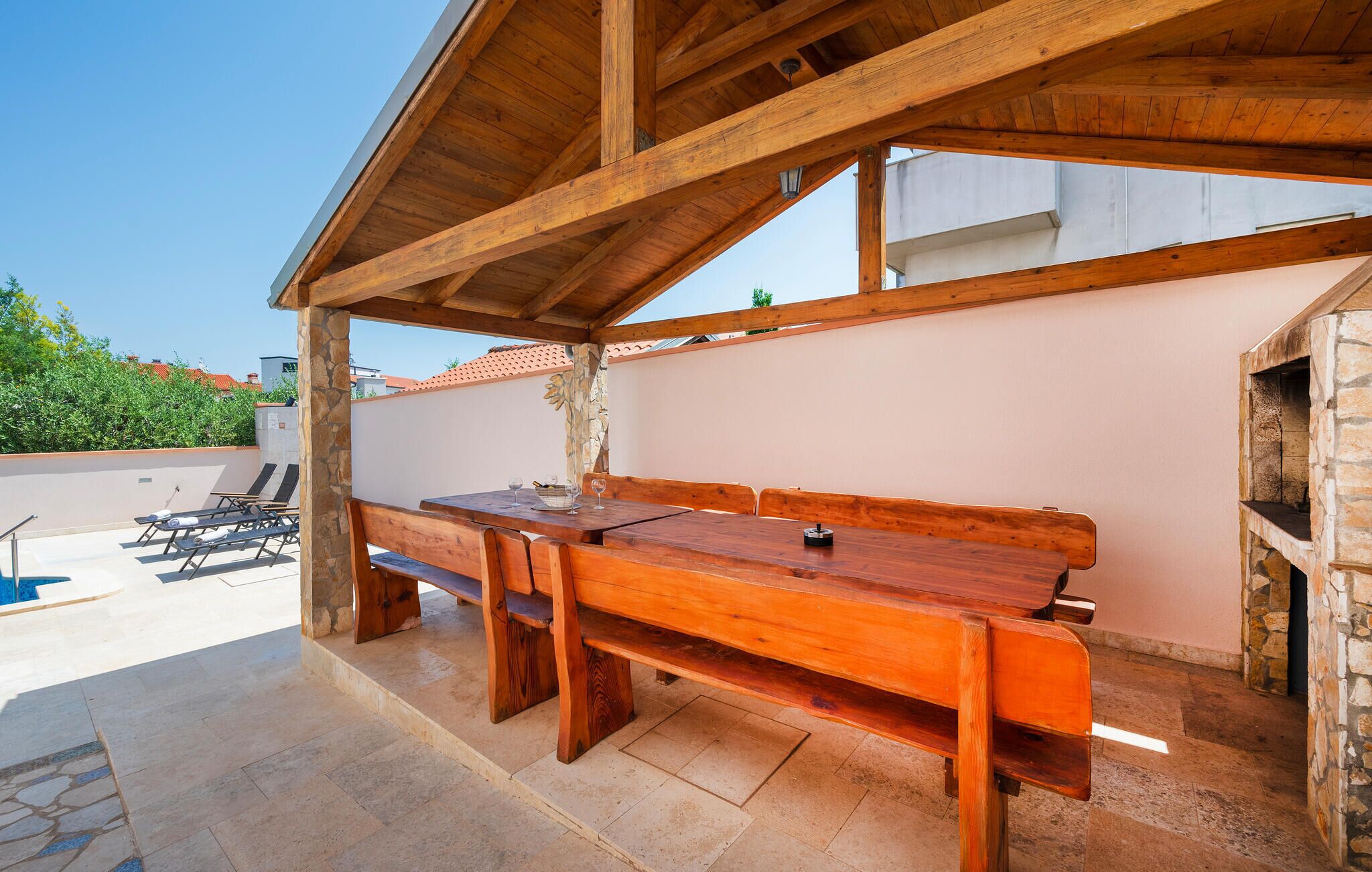 An outdoor patio with a wooden table and benches, stone walls, and a stone fireplace.