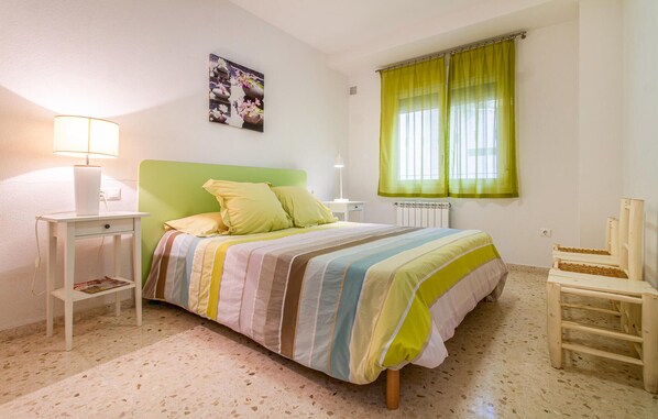A bedroom with a bed, a nightstand, a chair, a window with green curtains, and a wall-mounted picture.