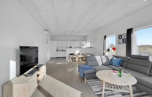 A modern living room with a sofa, a coffee table, and a TV. There is a kitchen in the background with white cabinets and a dining area.