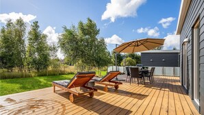 Eine Holzterrasse mit zwei Liegestühlen und einem Tisch, einem Grill und einem überdachten Bereich mit Sonnenschirm.