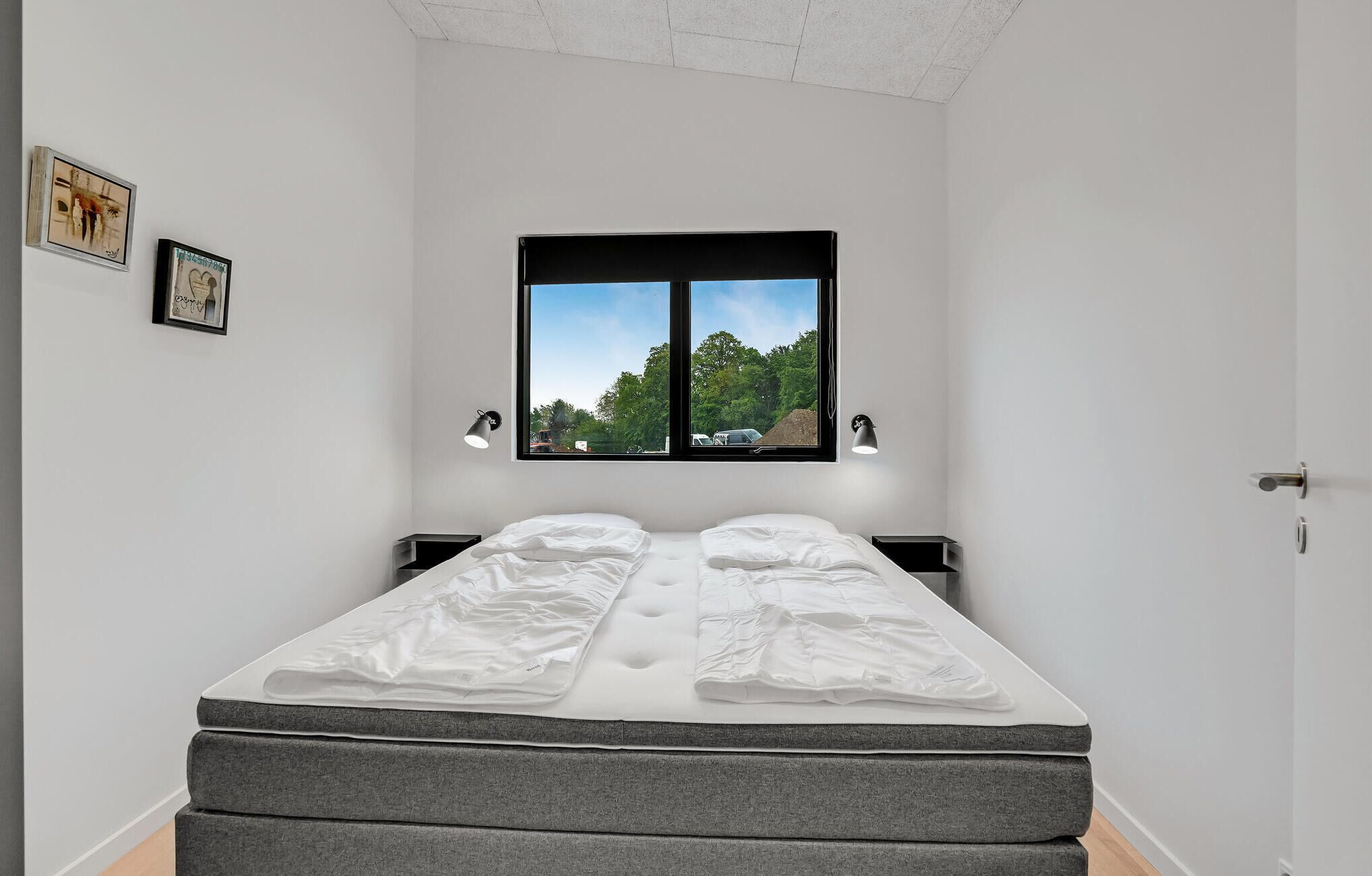 Un dormitorio con cama, dos mesitas de noche, ventana con vistas a árboles y dos lámparas de pared.