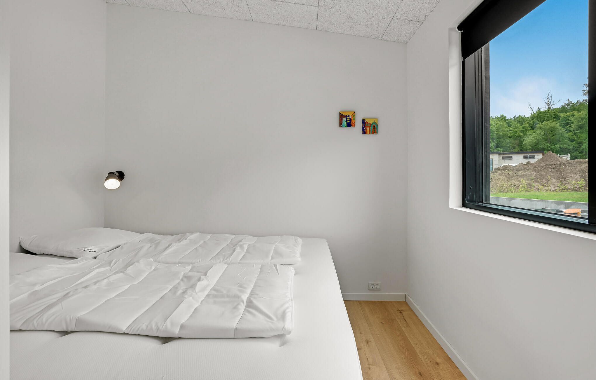 A bedroom with a bed, a nightstand, a wall-mounted lamp, and a window with a view of greenery and buildings.
