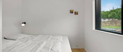 A bedroom with a bed, a nightstand, a wall-mounted lamp, and a window with a view of greenery and buildings.