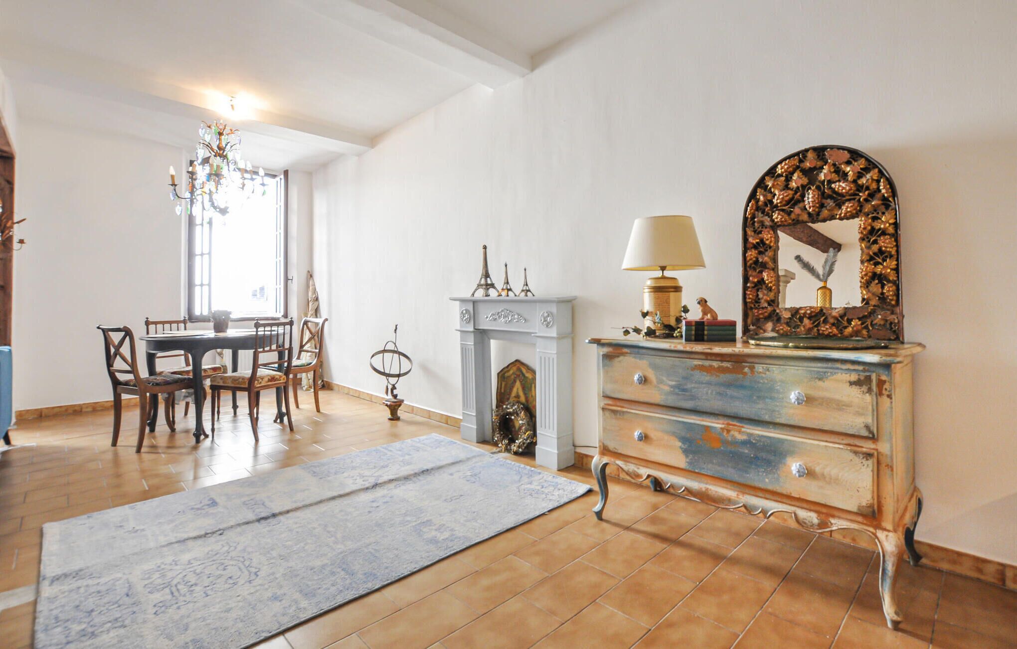 Une pièce comprenant une table à manger et des chaises, une cheminée agrémentée d’objets décoratifs, une commode avec un miroir et un lustre.