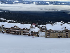 Un centro turístico cubierto de nieve con edificios de varios pisos, una pista de esquí y una montaña boscosa al fondo.