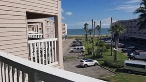 Uma varanda com vista para um estacionamento, uma praia e um edifício com palmeiras.