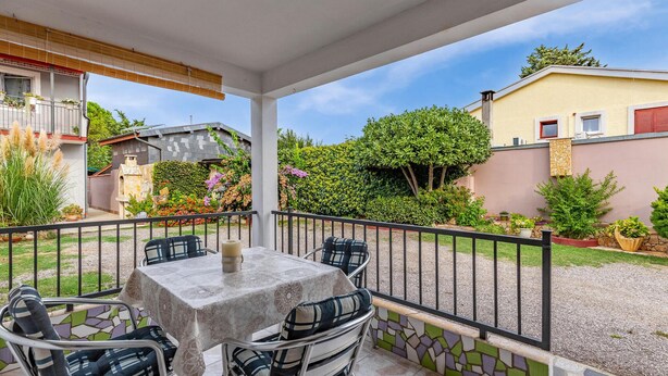 A covered patio with a table and chairs, surrounded by a black metal fence, and a garden with various plants and a house in the background. - 1 bedroom beach front apartment in Premantura (Premantura)