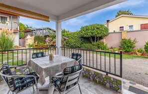 Une terrasse couverte avec une table et des chaises, entourée d’une clôture en métal noir, et un jardin avec différentes plantes, avec une maison en arrière-plan.