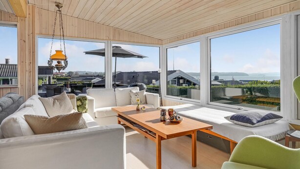 A modern living room with a large window, a wooden ceiling, and a view of the sea and buildings. - 4 bedroom cozy home in Rønde (Rønde)