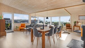 A spacious living area with a wooden ceiling, large windows offering a view of the sea, a dining table with chairs, and a sofa. - 4 bedroom cozy home in Rønde (Rønde)