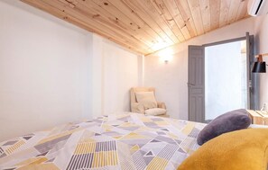 Une chambre avec un plafond en bois, un lit recouvert d’une couvre-lit à motifs géométriques, une chaise et une porte donnant sur une autre pièce.