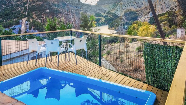 A pool area with a view of mountains and a river. There are white chairs and a table on a wooden deck.