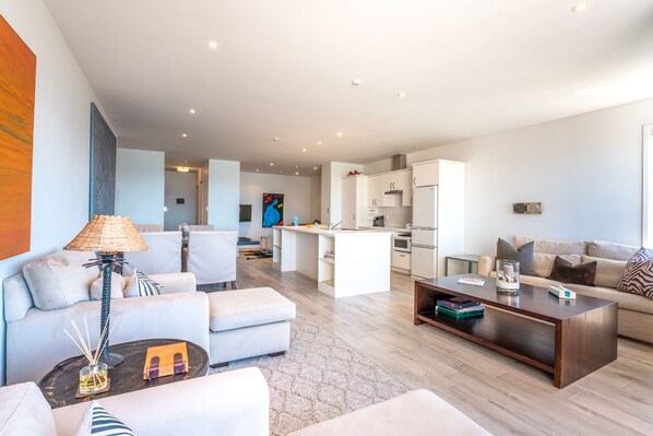 A modern living room with a sofa, coffee table, and a dining area with a kitchen in the background.