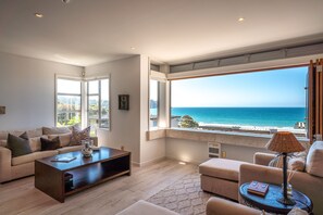 A modern living room with a large window overlooking the sea, a sofa, a coffee table, and a lamp. - Sunshine on The Beach at The Sands (Waiheke Island)